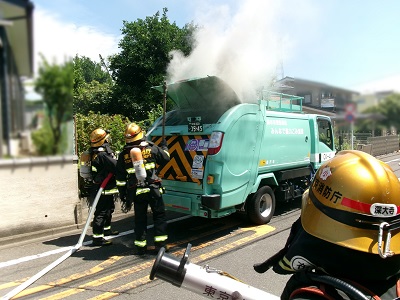 ごみ収集車両の後部ハッチから白煙が出ているのを確認する消防隊員の画像